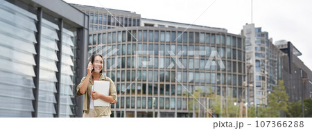 Stylish urban girl walking on street, talking on mobile phone and carrying laptop. Young woman making a telephone call while going somewhere 107366288
