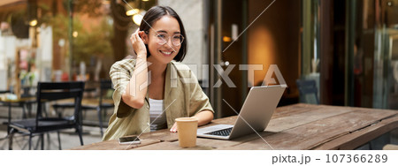 Portrait of smiling girl in glasses, sitting with laptop in outdoor cafe, drinking coffee and working remotely, studying online 107366289