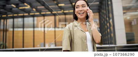 Happy asian woman talking on mobile phone, standing on bus stop in city. Outdoor shot 107366290