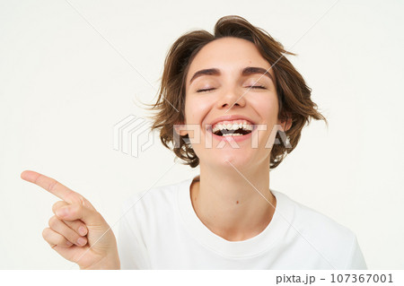 Close up portrait of brunette girl with happy face, laughing, pointing finger left, showing advertisement, standing over white studio background 107367001