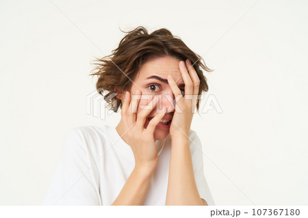 Close up of brunette woman hiding her face, screams and looks frightened, horrified by something, standing scared against white background Close up of brunette woman hiding her face, screams and looks frightened, horrified by something, standing scared against white background 107367180