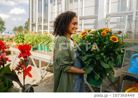 Smiling woman gardener holding flower pot while standing on greenhouse yard background 107368449