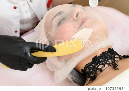 The beautician uses a spatula to apply the cream to the already prepared face with gauze. The beautician uses a spatula to apply the cream to the already prepared face with gauze. 107373984