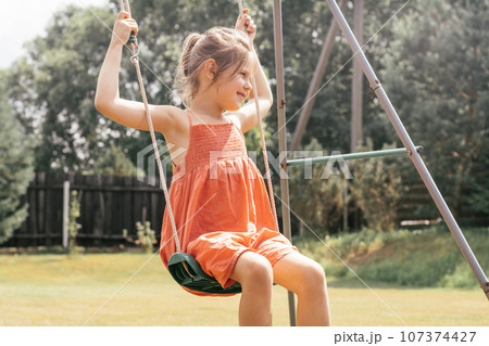 Happy little girl in orange sundress is carelessly swinging on swing on sunny summer day in nature. 107374427