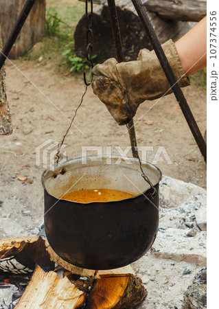 Hand of man in glove hanging cauldron with soup on hook of tripod over firewood, preparing for cooking on campfire. Hand of man in glove hanging cauldron with soup on hook of tripod over firewood, preparing for cooking on campfire. 107374556