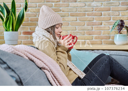 Young woman in sweater hat sitting at home, basking with cup of hot drink Young woman in sweater hat sitting at home, basking with cup of hot drink 107376234