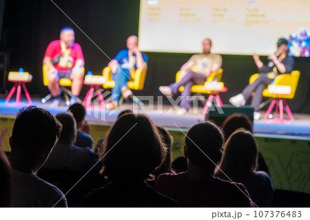 Colleagues sitting on stage during conference answering questions from audience in dark hall while presenting new project Colleagues sitting on stage during conference answering questions from audience in dark hall while presenting new project 107376483