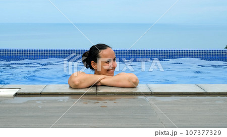 Happy young woman enjoying swimming in outdoor pool. Blue sea view on the background Happy young woman enjoying swimming in outdoor pool. Blue sea view on the background 107377329