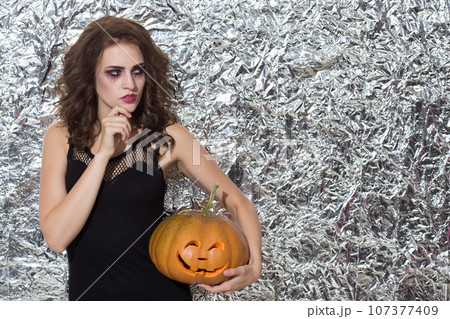 Pensive beautiful woman with decorative makeup, posing with pumpkin at Halloween. Front view of thoughtful female witch looking away, while plotting something, against foil wall. Concept of Halloween. 107377409