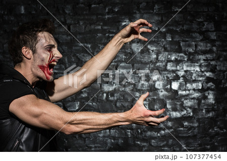 Waist up photo of a young man with bloody make up, standing in a profile shouting and stretching his hands to copy place on the black brick wall Waist up photo of a young man with bloody make up, standing in a profile shouting and stretching his hands to copy place on the black brick wall 107377454