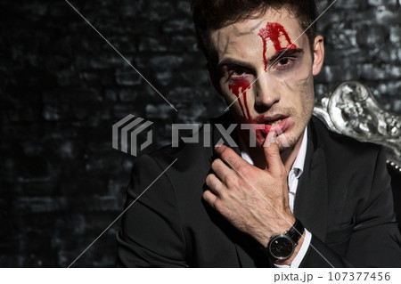 Portrait of a handsome young man sitting on a couch wearing a suit, wiping blood from his lip with his finger Portrait of a handsome young man sitting on a couch wearing a suit, wiping blood from his lip with his finger 107377456