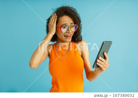 Shoked woman holding his head while looking on mobile phone over blue studio background 107380032