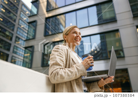 Woman manager drinking coffee while working laptop on modern building background and looks away 107380384