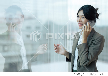 Smiling businesswoman looking through the window while talking on mobile phone in the office. Copy space 107380746