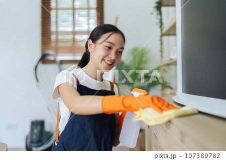 Young woman cleaning using spray and wiping the cabinet tv with microfiber cloth in living room 107380782
