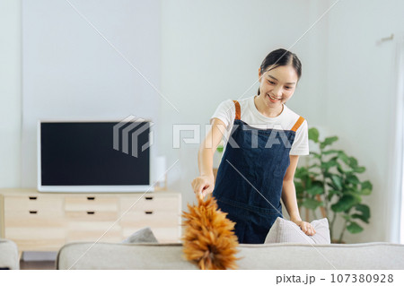 young female housekeeper taking up pillow dusting the couch in the cozy living room 107380928