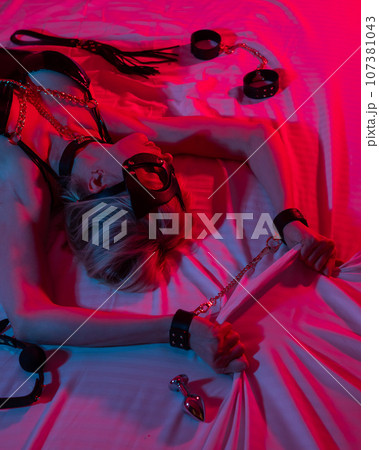 Sexy blonde woman in a leather mask and handcuffs and a collar lies in the bedroom in a red-blue neon light. Role-playing games. Sexy blonde woman in a leather mask and handcuffs and a collar lies in the bedroom in a red-blue neon light. Role-playing games. 107381043