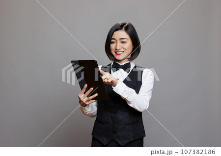 Smiling restaurant receptionist in uniform checking guest list on digital tablet. Eatery asian woman waitress tapping on touchscreen while examining online order while posing in studio 107382186