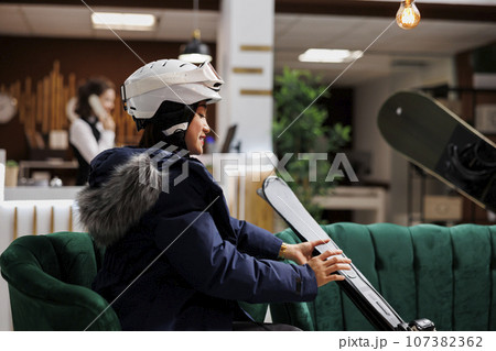 Asian female traveler in winter jacket comfortably seated on sofa with her ski equipment in hotel reception. Enthusiastic woman with snow helmet placing her skiing skis nearby in lounge area. Asian female traveler in winter jacket comfortably seated on sofa with her ski equipment in hotel reception. Enthusiastic woman with snow helmet placing her skiing skis nearby in lounge area. 107382362