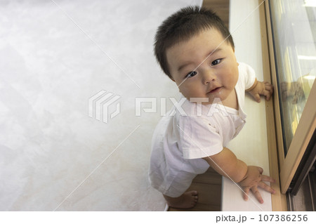 窓際でつかまり立ちするカメラ目線の赤ちゃん（男の子、生後9カ月、1歳、日本人） 107386256