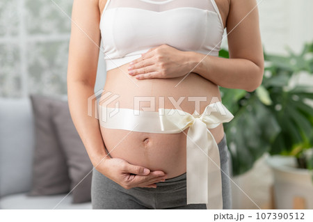 Close up Pregnant Woman standing in front of windows and stroking her big belly with love at cozy home,Pregnancy of young woman enjoy with future life relax with love,Motherhood and Pregnant Concept 107390512