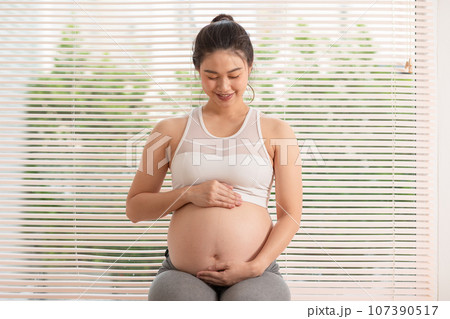 Happy Pregnant Asian Woman sitting on a bed holding and stroking her big belly at home. Pregnancy of young woman enjoying with future life relax at home. Motherhood and Pregnant Concept. Soft focus 107390517