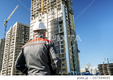 Builder at a construction site. Construction of a high-rise building and a builder looking at the process Builder at a construction site. Construction of a high-rise building and a builder looking at the process 107394695