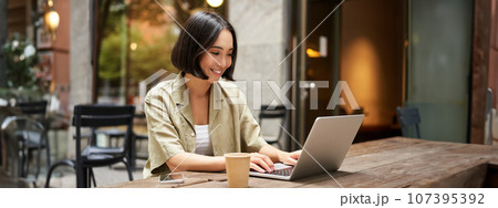 Young asian woman, digital nomad working remotely from a cafe, drinking coffee and using laptop, smiling Young asian woman, digital nomad working remotely from a cafe, drinking coffee and using laptop, smiling 107395392