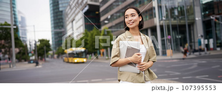 Beautiful asian girl smiles as commutes to work, stands on street with laptop and notebook Beautiful asian girl smiles as commutes to work, stands on street with laptop and notebook 107395553