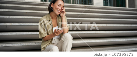 Portrait of happy asian girl sits with mobile phone and laughs. Young woman with smartphone being outside 107395575