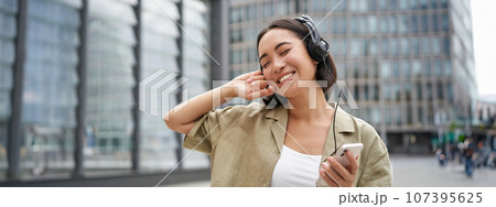 Happy young woman dancing on streets and listening music in headphones, holding smartphone 107395625