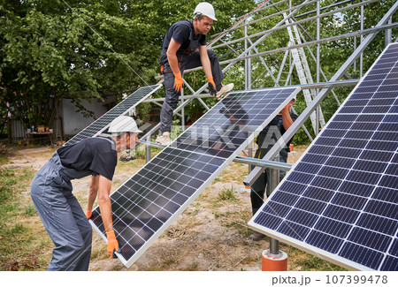 Three men solar installers wearing safety helmets and work overalls while mounting photovoltaic solar panel system. Male workers installing solar module on metal construction. 107399478