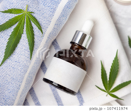 Dropper bottle with blank label near green cannabis leaves top view, CBD cosmetic mockup 107411974