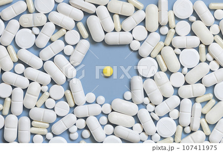 Mix of white medical capsules and pills with one yellow pill in the middle on light blue top view 107411975
