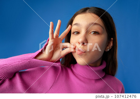 Portrait of teen girl showing silence gesture over blue background 107412476
