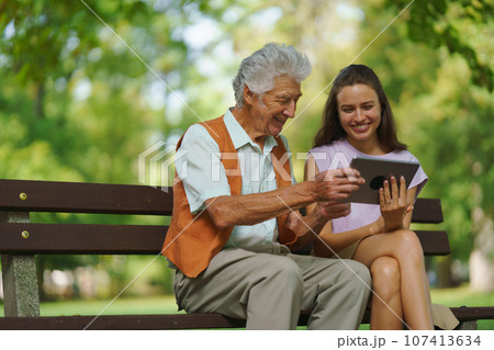 The grandfather and granddaughter are looking at photos on a tablet, sitting on a bench in a city park. 107413634