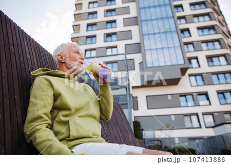 Elderly man sitting on bench, drinking water after his morning run. 107413886