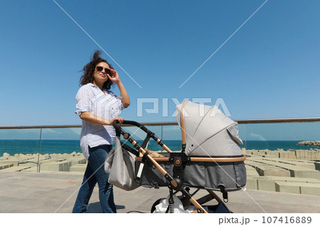Beautiful woman enjoying a promenade along Atlantic seacoast in warm sunny day, pushing a stroller with her newborn baby 107416889