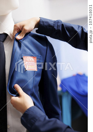 Male customer checking discount amount looking at price on formal jacket while buying clothes on Black Friday, cropped photo. Retail worker sticking red sale tag on clothing, working in outlet store 107418031