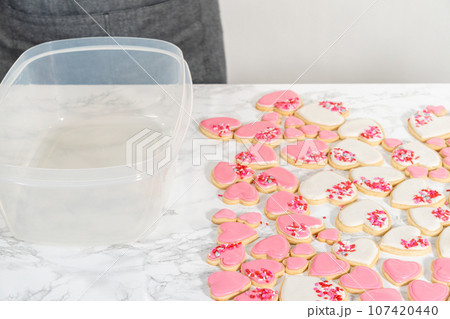 Storing heart-shaped sugar cookies with pink and white royal icing in a large plastic container. Storing heart-shaped sugar cookies with pink and white royal icing in a large plastic container. 107420440