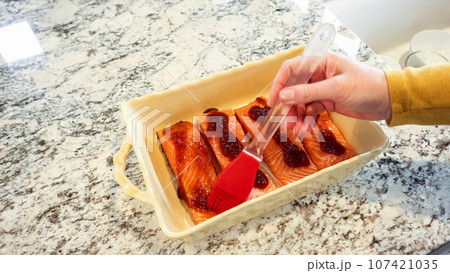 In a modern white kitchen, the raw salmon is being generously covered with teriyaki sauce. 107421035