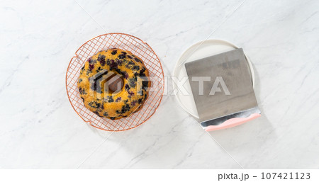 Flat lay. Transferring cooled freshly baked lemon blueberry bundt cake from the round cooling rack to the serving plate. Flat lay. Transferring cooled freshly baked lemon blueberry bundt cake from the round cooling rack to the serving plate. 107421123