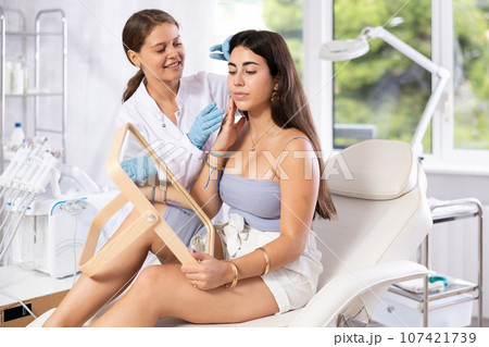 Cosmetologist explaining facial procedure to young girl looking in mirror 107421739