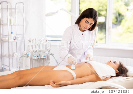 Cosmetologist performing ultrasound cavitation procedure on belly for female client 107421883