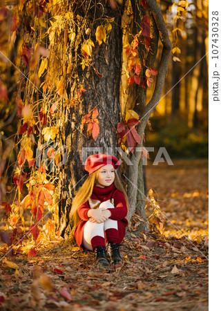 Child girl is sitting near a tree in an autumn city park, a child is playing and enjoying an autumn day 107430328