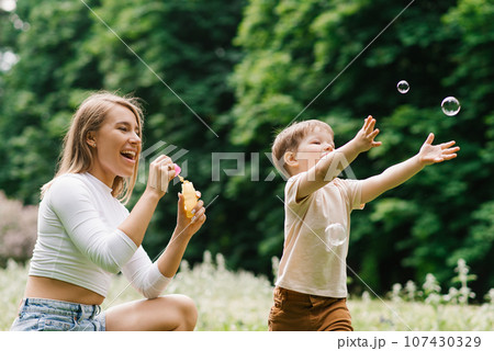 Young Caucasian mother and her happy little son are having fun playing with soap bubbles in the park Young Caucasian mother and her happy little son are having fun playing with soap bubbles in the park 107430329