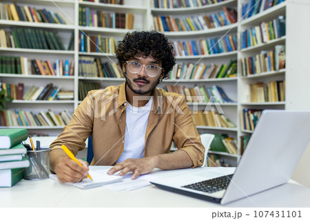 Portrait of a young Indian male student writing an outline while sitting in a university library at a table with a laptop. Portrait of a young Indian male student writing an outline while sitting in a university library at a table with a laptop. 107431101