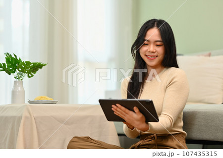 Happy young woman sitting living room checking social network, watching video on digital tablet Happy young woman sitting living room checking social network, watching video on digital tablet 107435315