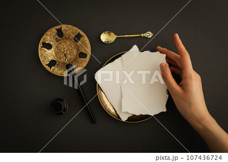 Papers, female hand, golden plates and spoon, ink bottle and pen on dark gray background, top view 107436724