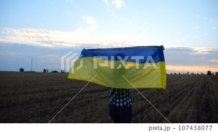 Ukrainian lady walking on barley meadow with raised over head blue-yellow banner at sunset. Woman going on wheat field with flag of Ukraine at sunrise. Victory against russian aggression. 107438077